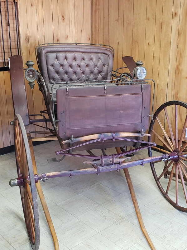 An antique wooden carriage with a tufted purple cushion seat and a storage chest on the front. The carriage has large spoke wheels and metal accents, giving it a classic, rustic appearance.