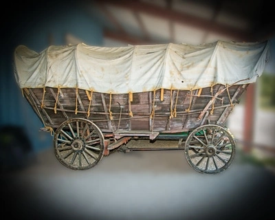 An old-fashioned covered wagon with a wooden frame and canvas top. The wagon is on large wooden wheels and appears to be in a rustic, outdoor setting.