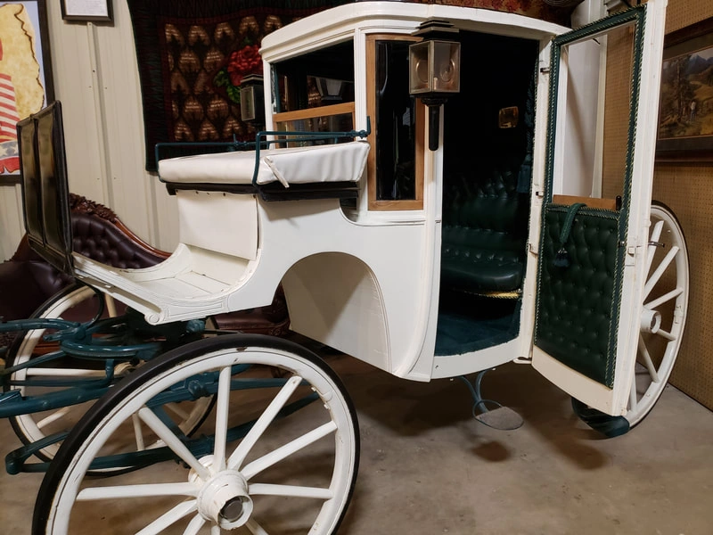An antique white horse-drawn carriage with large wooden spoked wheels and a curved glass window visible on the side.