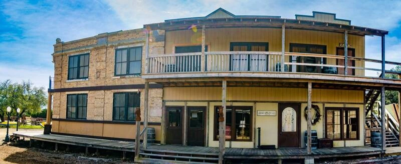 A two-story, tan-colored building with a wraparound porch and large windows. The building has a balcony on the second floor and a sign above the front entrance. The building appears to be in a rural or small-town setting, with some trees and a clear blue sky in the background.
