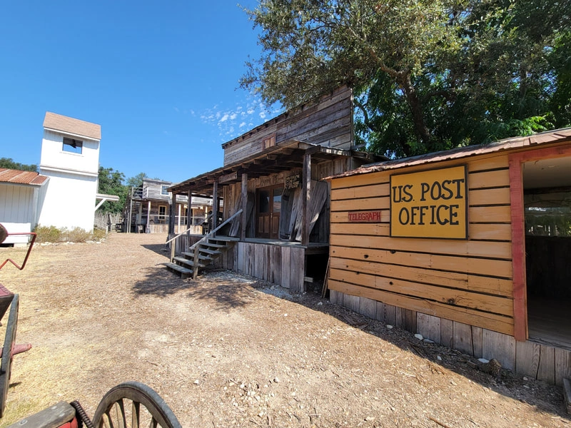 A rustic wooden building with a sign that says "US POST OFFICE". There are other smaller wooden structures in the background, and the scene is surrounded by trees and bushes.