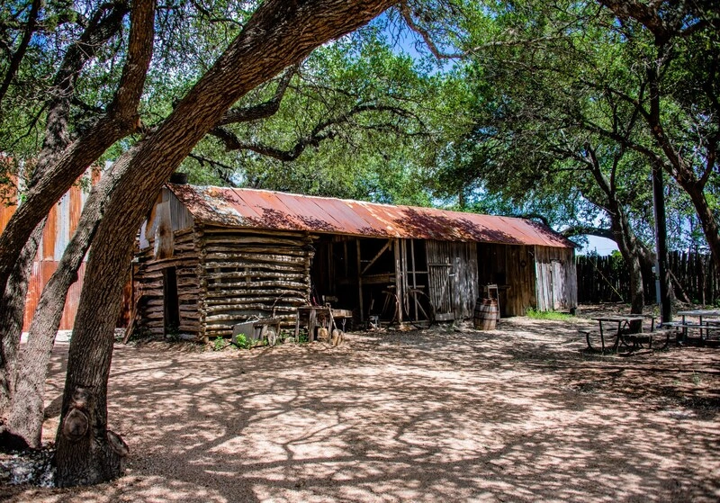 An old wooden barn or ranch structure with a red tin roof, surrounded by lush trees and greenery. The building appears weathered and rustic, sitting on a sandy dirt path.