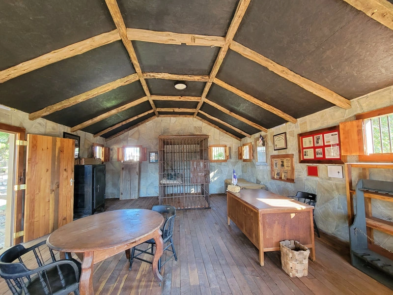 The image shows the interior of a rustic, wooden cabin or shed. The space has a high, pitched ceiling with exposed wooden beams and trusses. There is a large wooden table in the center of the room, surrounded by wooden chairs. The walls are made of wooden planks, and there are various items and furnishings scattered throughout the space, giving it a lived-in, cozy feel.