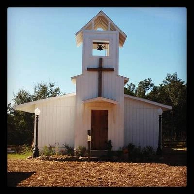 A white wooden church building with a bell tower and a cross on top, surrounded by trees and a grassy area.