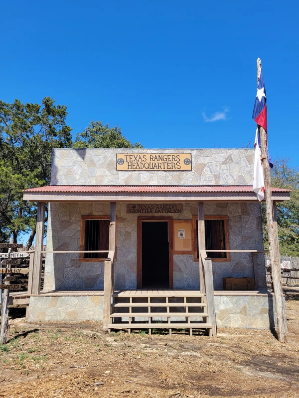 A rustic wooden building with the Texas Ranger sign displayed, set against a clear blue sky and surrounded by trees.