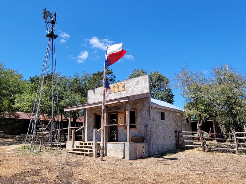 An old Western-style wooden building with a windmill and Texas flag on a sunny day