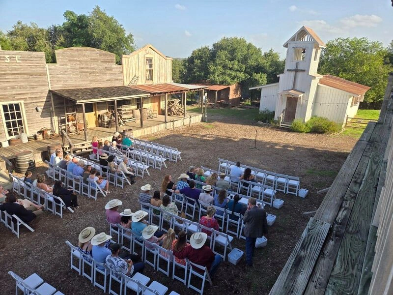 An outdoor wedding ceremony setup with rows of white chairs facing a small raised platform with a decorated archway. Several large trees and buildings are visible in the background, creating a rustic, countryside setting.