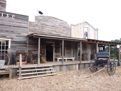 A rustic, wooden Western-style building with a covered porch and a horse-drawn carriage parked in front. The building has an old, weathered appearance, typical of a historic frontier town setting.