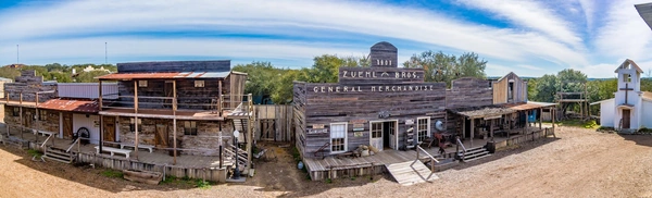 Panoramic view of an old western style town with wooden buildings, storefronts, and a water tower in the background.