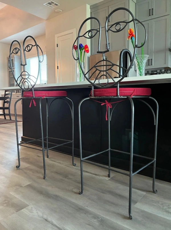 Two ornate metal bar stools with circular backrests and red upholstered seats sit at a kitchen island in a bright, modern home. The stools feature decorative scrollwork and ribbon accents, set on a light wood floor with gray cabinetry and a white ceiling. This interior furniture shot aligns with items Burgess Estate Sales may curate for estate sales in Central Texas.