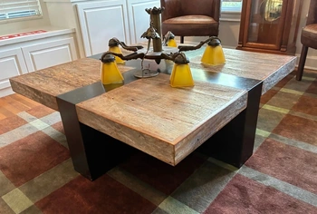 A unique wooden coffee table with a chandelier centerpiece featuring yellow glass shades, displayed on a plaid rug in a room with white paneled walls and a brown leather armchair.