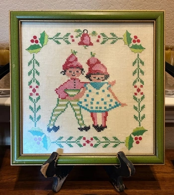 Framed cross-stitched artwork featuring two children in vintage winter attire, surrounded by holly and bells, displayed on a stand.