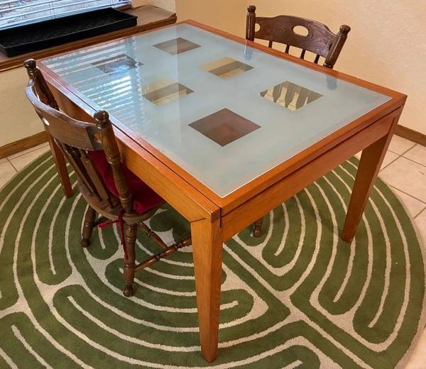 A square wooden dining table with a glass top and decorative inset panels, flanked by two wooden chairs, sitting on a green circular-pattern rug-a sample of vintage furniture often showcased in Burgess Estate Sales’ Austin-area estate liquidations.