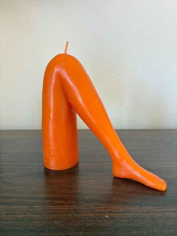 Orange candle sculpted into a bent leg with a high-heeled foot, perched on a dark wooden surface against a pale wall. A quirky, modern décor piece-great for estate sale listings and vintage home decor in Austin and Central Texas.
