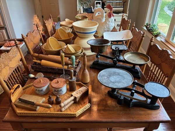 Vintage kitchen display featuring antique balance scales, yellowware mixing bowls, decorative ceramic rooster, carved wooden chairs, and various wooden kitchen tools including rolling pins and spice containers on a wooden table