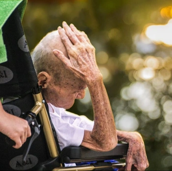 An elderly man in a wheelchair appears to be overwhelmed or distressed, with his hands covering his face. The image has a somber, emotional tone and suggests the challenges faced by those with mobility issues or in need of care.