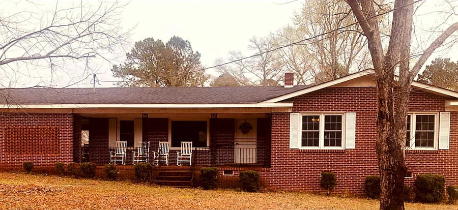 Classic red brick ranch-style home with covered front porch featuring white rocking chairs, decorative shutters, and well-maintained shrubs surrounded by fallen autumn leaves
