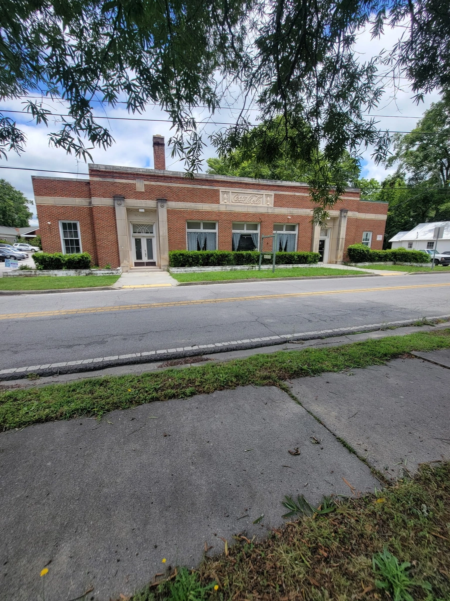 Historic red brick Coca-Cola bottling facility with Art Deco architectural elements, featuring limestone accents, large windows with navy curtains, and a prominent chimney, surrounded by manicured hedges and tree-lined street