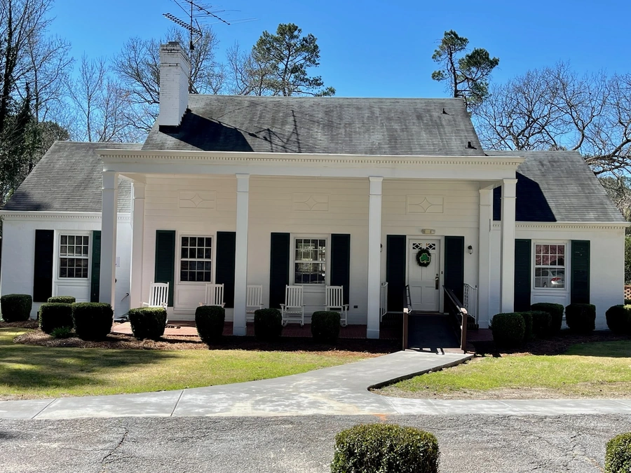 Traditional white Colonial-style house with a symmetrical facade, featuring a covered front porch with white columns, black shutters, rocking chairs, and manicured boxwood shrubs along the front