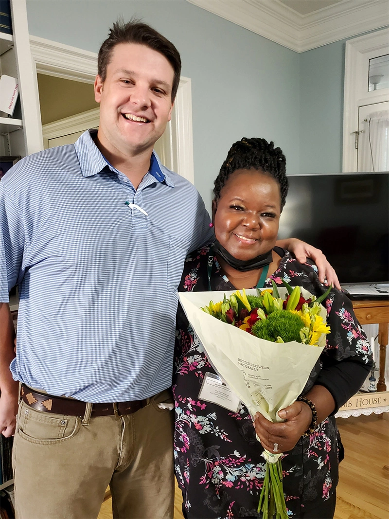 Two smiling colleagues standing together indoors, one wearing a blue striped polo shirt and khakis, the other in a black floral dress holding a vibrant bouquet of yellow sunflowers and mixed flowers wrapped in white paper