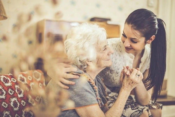 A heartwarming moment between a caregiver and elderly woman, showing them holding hands and sharing a gentle moment in a home setting with warm lighting