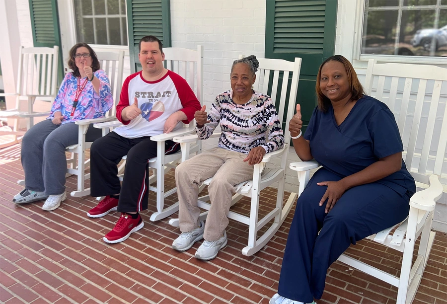 Four people sitting in white rocking chairs on a brick porch, including healthcare staff and residents giving thumbs up, with white painted building and green shutters in background