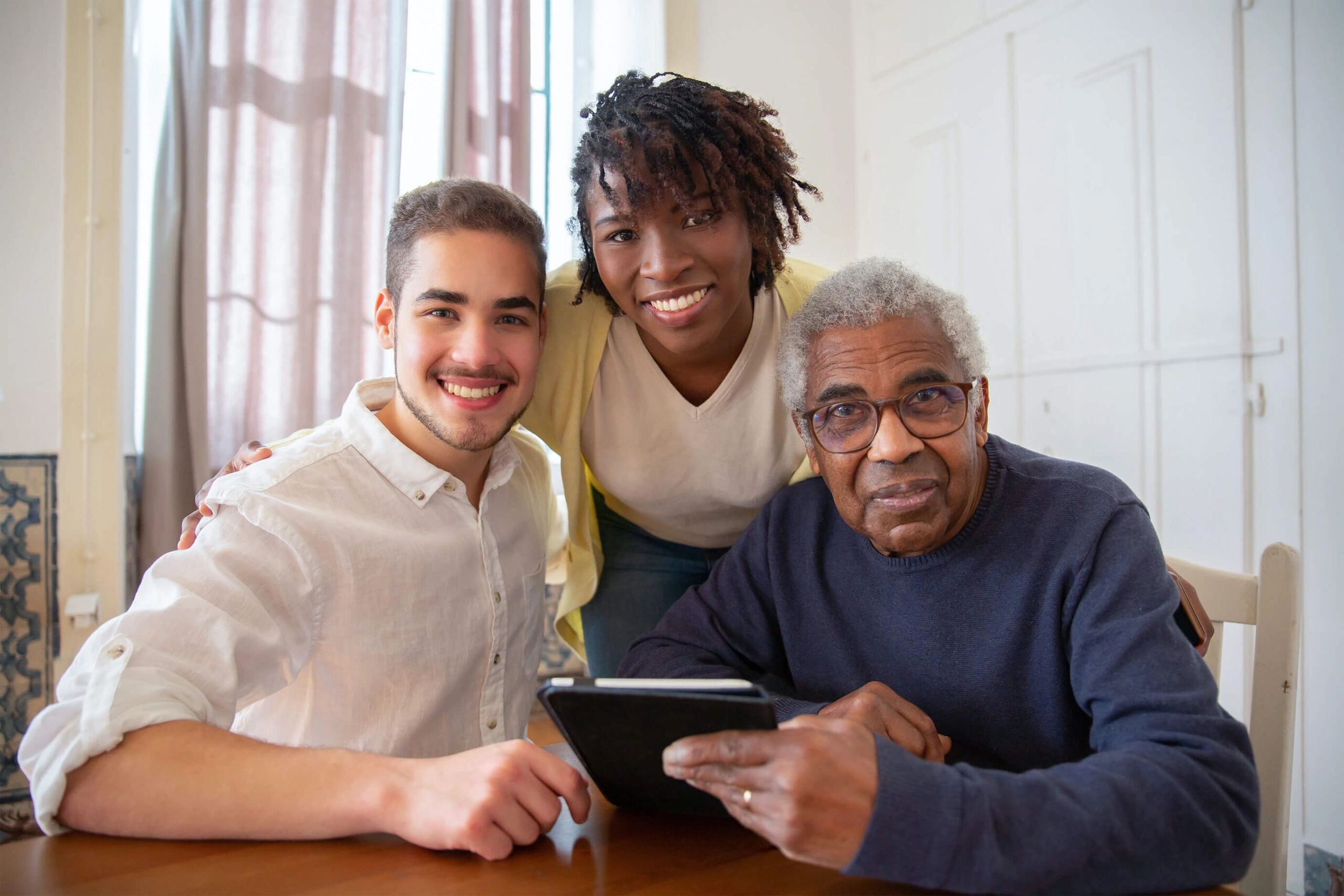 Three people of different generations gathered around a tablet device, smiling and engaging in what appears to be a technology learning session. The group includes a young adult in a white shirt, a middle-aged person in yellow, and a senior in a navy sweater, all looking engaged and happy while sharing the digital experience.