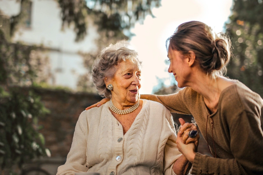 Heartwarming moment between an elderly woman in a cream cable-knit sweater and pearl necklace sharing a genuine connection with a younger caregiver in a brown sweater, both smiling warmly outdoors