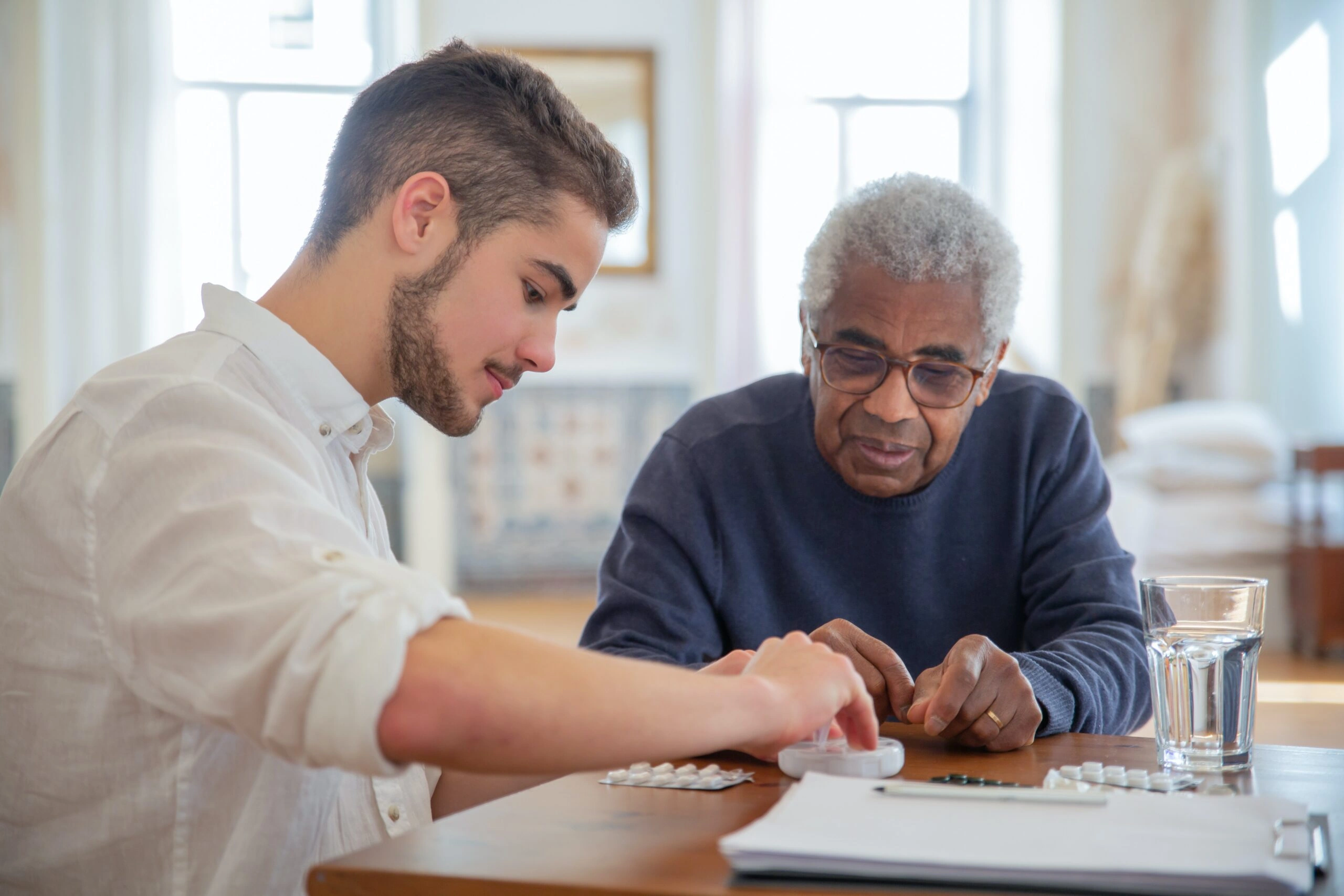 A younger man in a white shirt helping an older gentleman in a navy sweater organize medications at a table, with a glass of water and pill organizer visible
