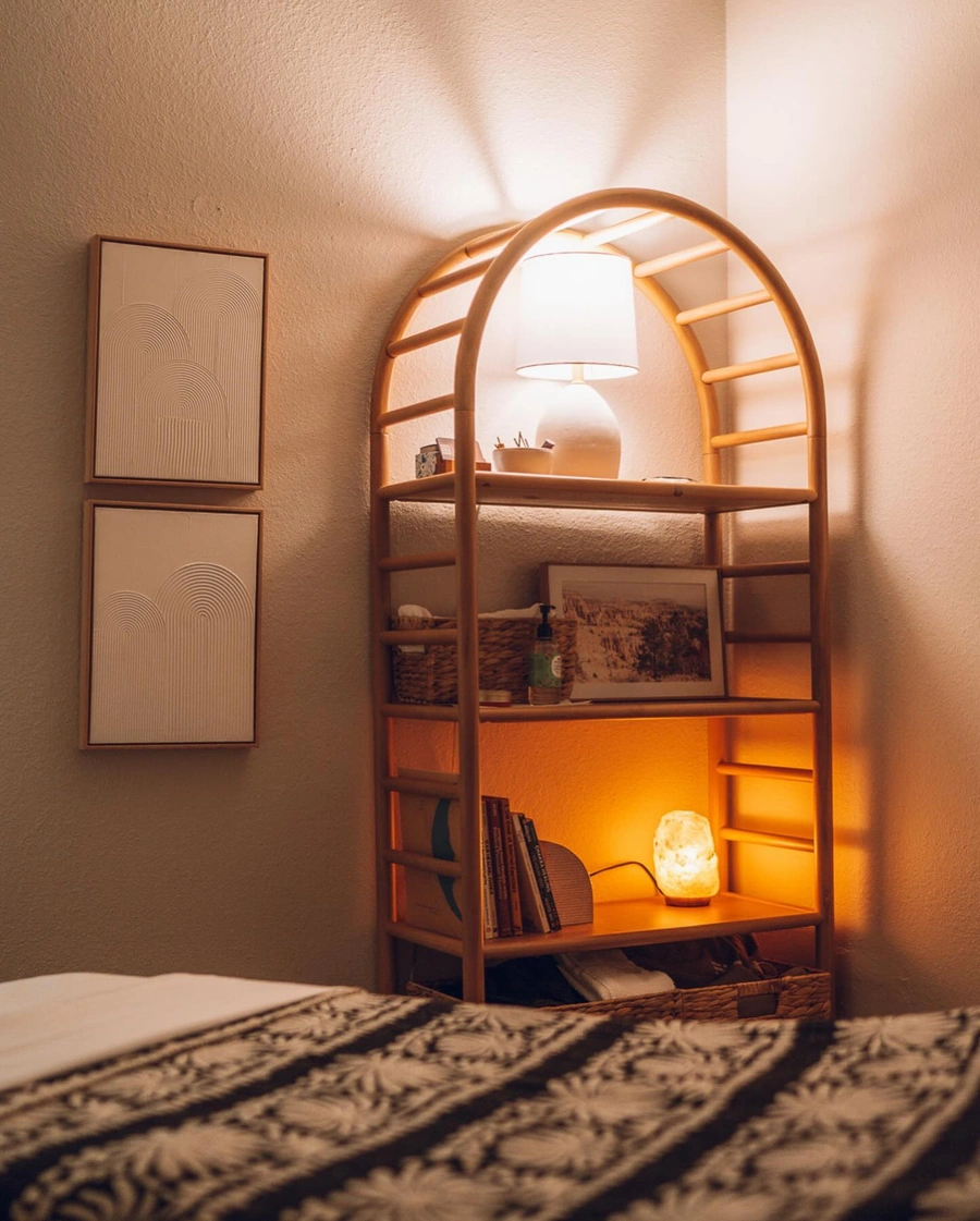 A cozy, warm-lit bedroom with a wooden shelving unit displaying books, plants, and decor items. The room has a wooden bed frame and patterned bedding, creating a peaceful and inviting atmosphere.