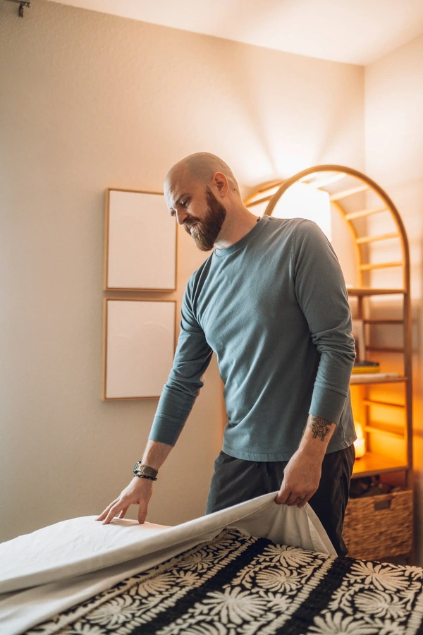 A man with a beard and tattoos stands in a room, leaning over a massage table with a decorative patterned blanket.