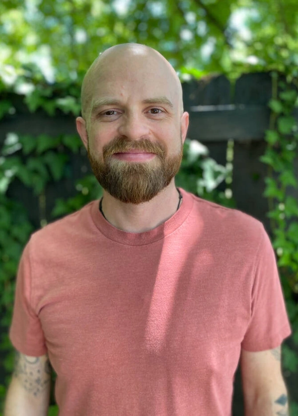 Portrait of a smiling man with a beard wearing a pink shirt against a blurred green background