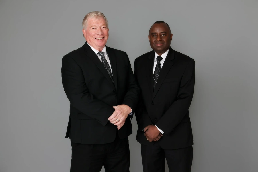 Firm partners in dark suits smiling against a gray studio backdrop; one older with light hair and a patterned tie.
