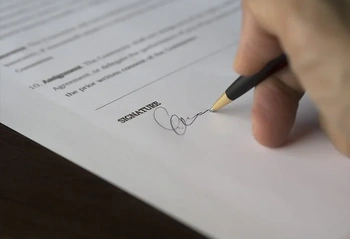 Close-up of a hand signing a legal document at a desk with the 'SIGNATURE' line visible