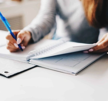 Person writing in a spiral notebook amid legal papers and a binder on an office desk
