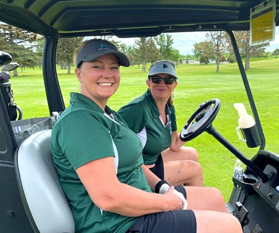 Two women in green polos seated in a golf cart on a golf course, one holding a water bottle