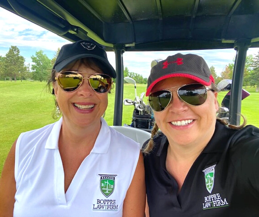 Two women in a golf cart wearing caps and sunglasses with golf clubs behind them and Boppre Law Firm logos on shirts