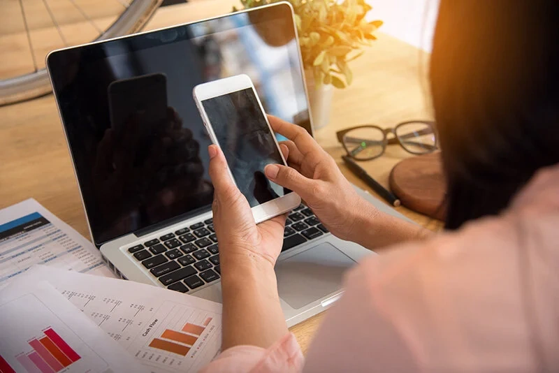 Professional at a wooden desk using a smartphone and laptop amid charts and documents