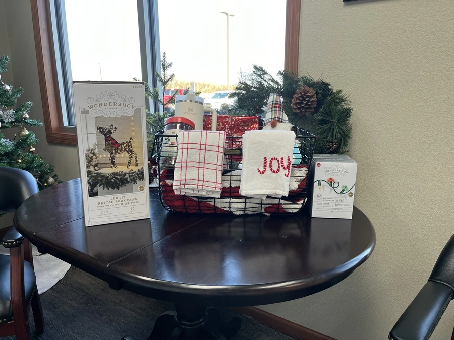 Holiday giveaway basket with 'JOY!' towels, gift tins, garland and a small decorated Christmas tree on a wood table