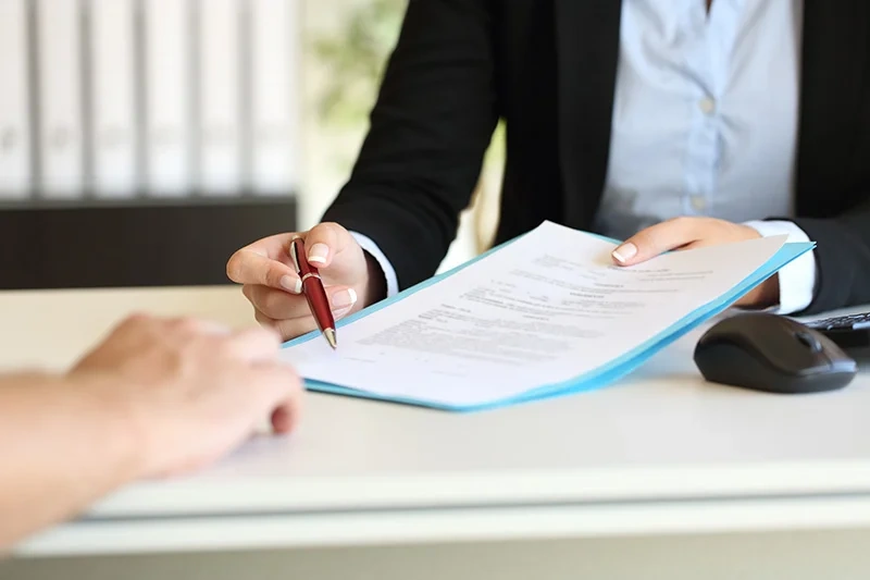 Two people at an office desk reviewing documents in a blue folder with a red pen - contract review meeting