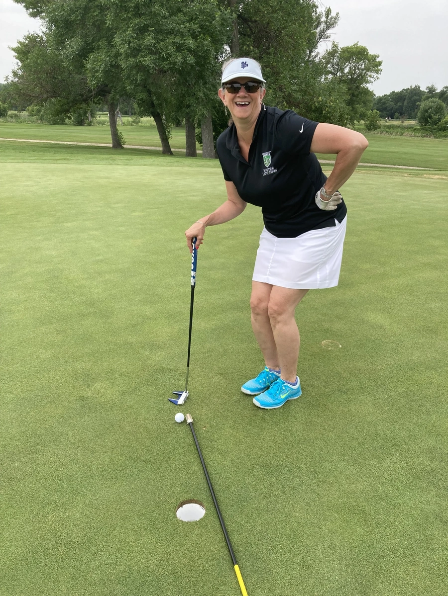 Woman smiling on a putting green holding a putter beside a golf hole with an alignment rod on the grass