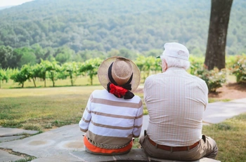 Elderly couple seated on a stone bench overlooking vineyard rows and distant hills
