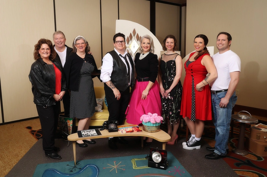 Nine people posing at a community gala in a room with retro decor and a yellow couch