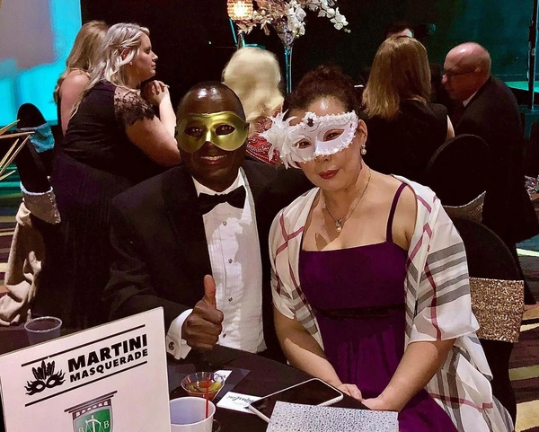 Two attendees wearing ornate masquerade masks at a 'Martini Masquerade' table; man in tuxedo giving a thumbs-up.