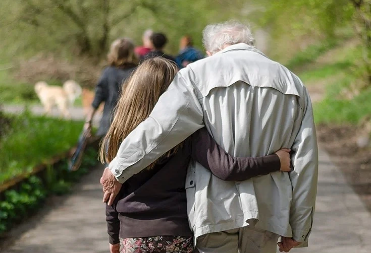 Elderly man walking arm-in-arm with younger woman along a tree-lined park path, suggesting family care