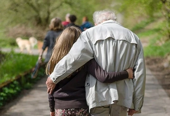 Elderly man walking arm-in-arm with younger woman along a tree-lined park path, suggesting family care