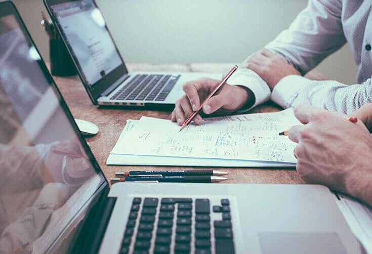Two people at a wooden desk using laptops and taking notes in a notebook, suggesting an estate-planning meeting