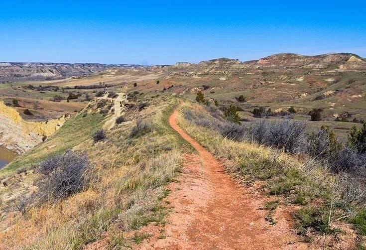 Red dirt trail winding along a grassy ridge with layered hills under a clear blue sky.