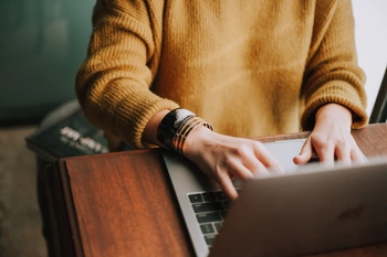 Person in a mustard sweater typing on a silver laptop at a wooden desk, wearing metal bracelets.