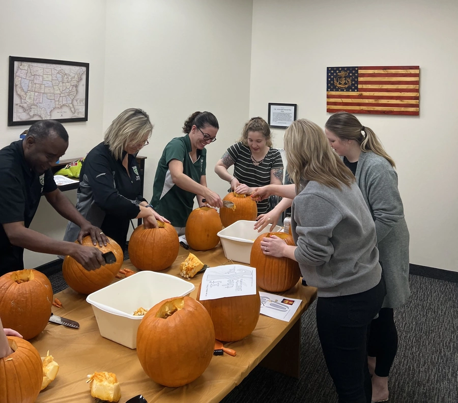 Community members carving pumpkins at a long table, using knives and spoons with bins for scraps.